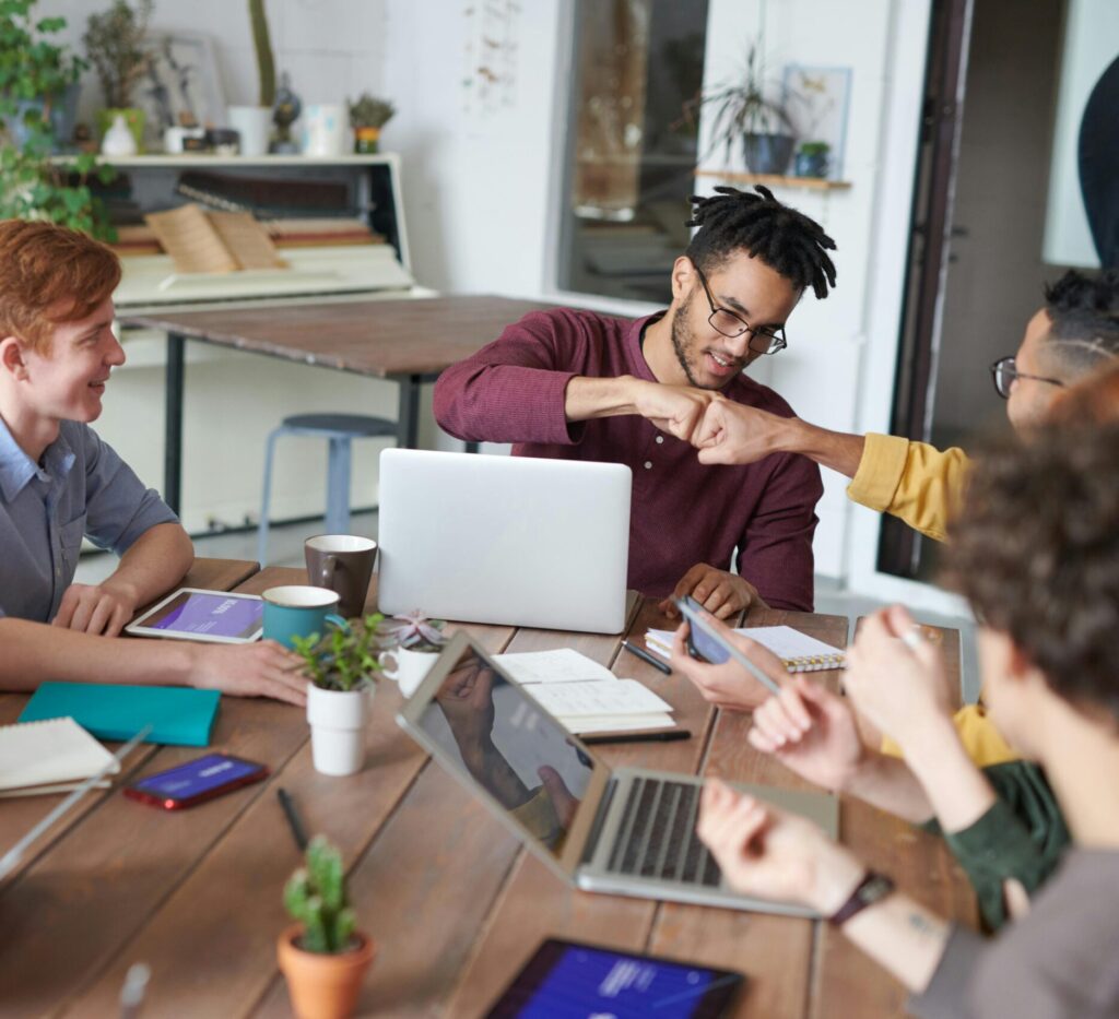 A group of young professionals collaborating at a table with laptops and notebooks. Two individuals exchange a fist bump, while others smile and engage in discussion.