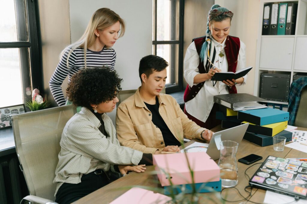 A diverse group of four people gathered around a table, collaborating on a project, with one person working on a laptop and others reviewing notes and files.