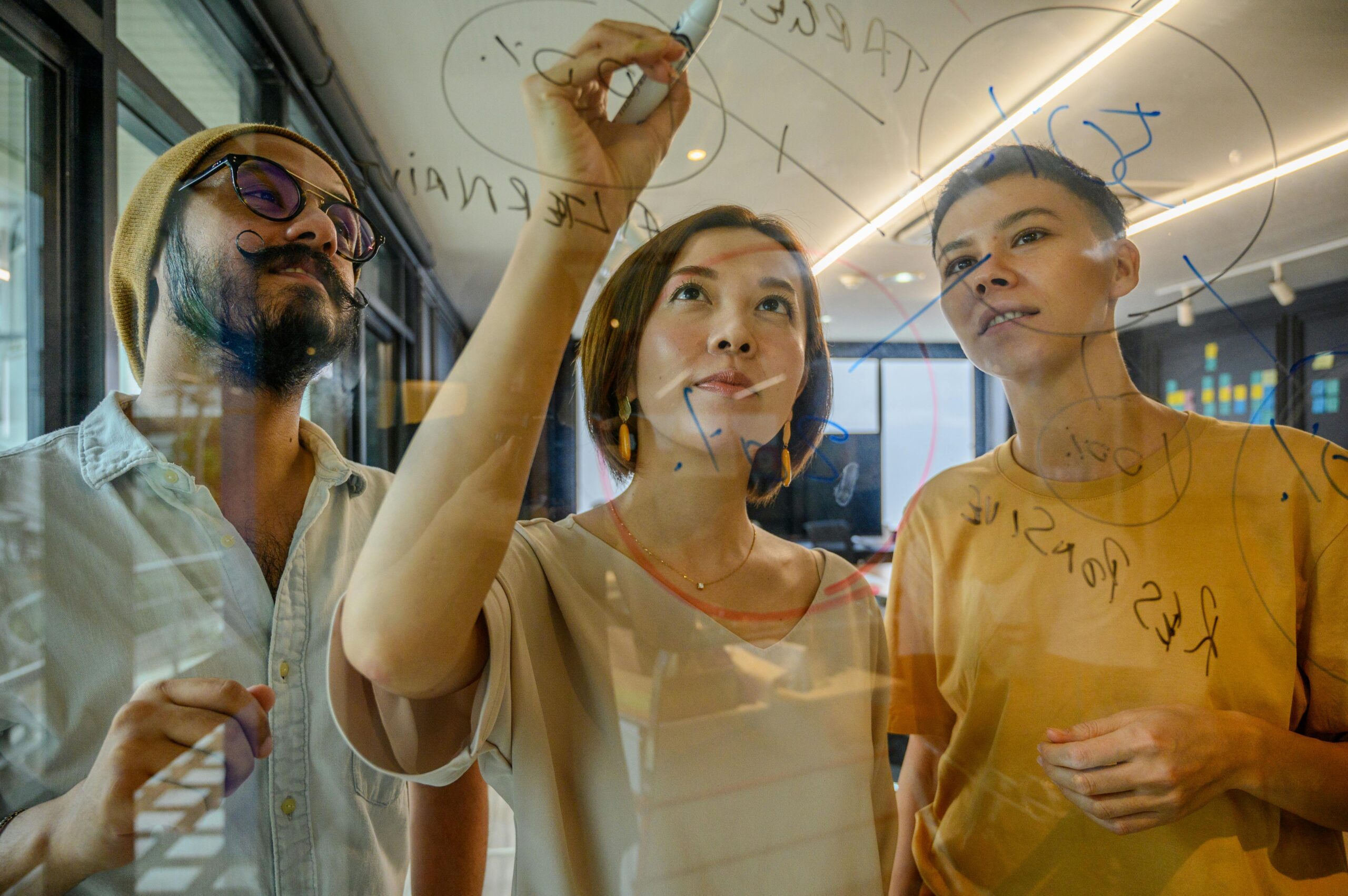 Three focused colleagues, two women and a man, writing and drawing on a transparent glass board in a modern office space during a brainstorming session.