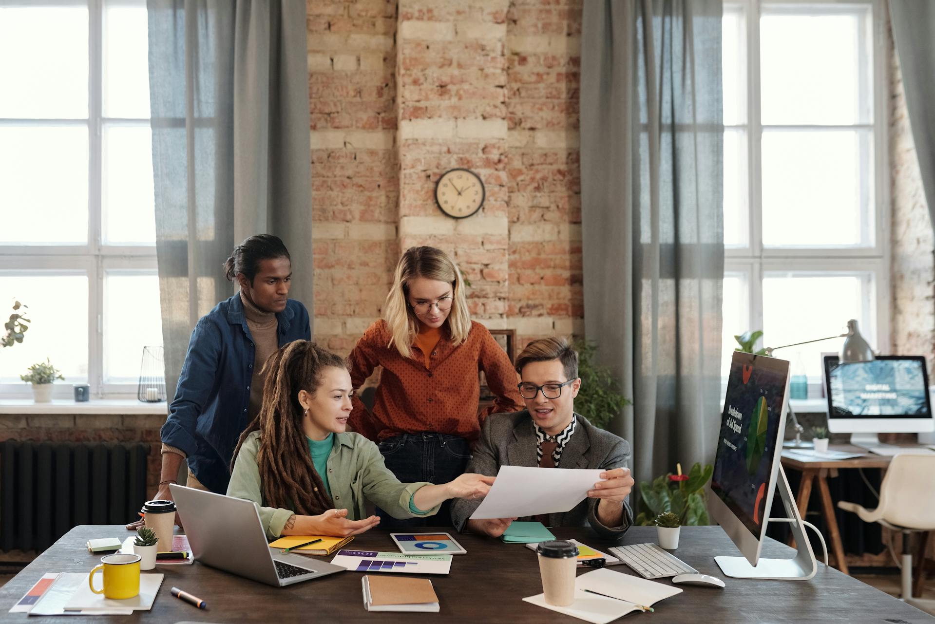 A diverse group of four colleagues collaborating at a wooden table in an industrial-style office with exposed brick walls and large windows.