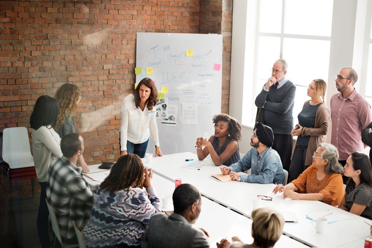 A diverse group of individuals gathered in a modern office, engaged in a brainstorming session led by a woman standing at the front near a whiteboard.