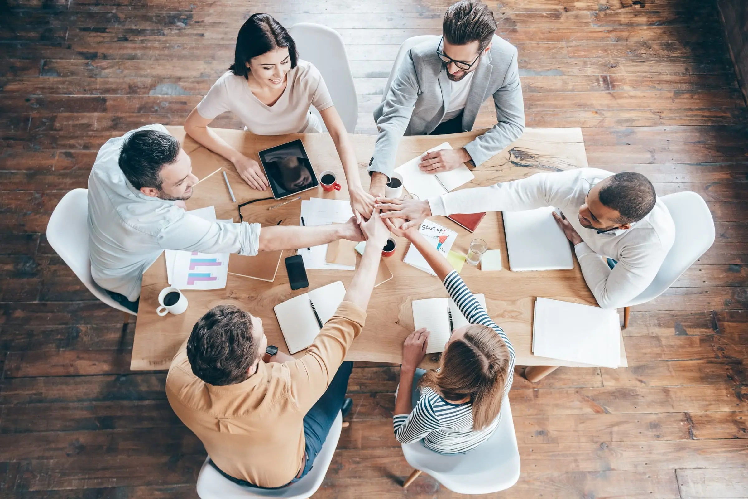 Top-down view of a diverse team sitting around a table, joining hands in a show of unity and teamwork.